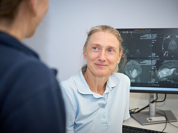 Hier sieht man Dr. Annette Wessmann im Gespräch mit einer Besitzerin. Im Hintergrund ist ein Bildschirm zu sehen, der MRT-Bilder zeigt.