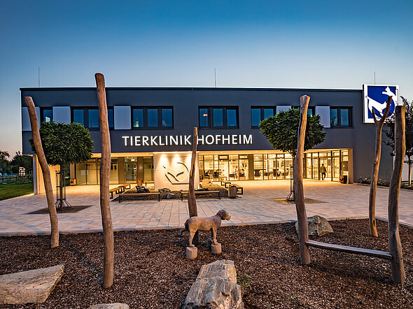Dieses Bild zeigt eine Außenansicht der Tierklinik Hofheim in der Abenddämmerung. Der Eingangsbereich ist beleuchtet. Im Vordergrund sieht man Baumstämme, die als Hundetoilette dienen. 