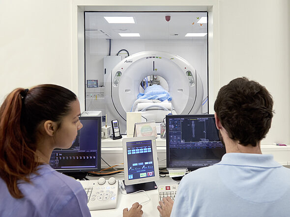Hier liegt ein Patient im CT unter einer wärmenden Decke. Ein Tierarzt und eine TFA beobachten ihn durch ein Fenster und steuern das Gerät von außen. 