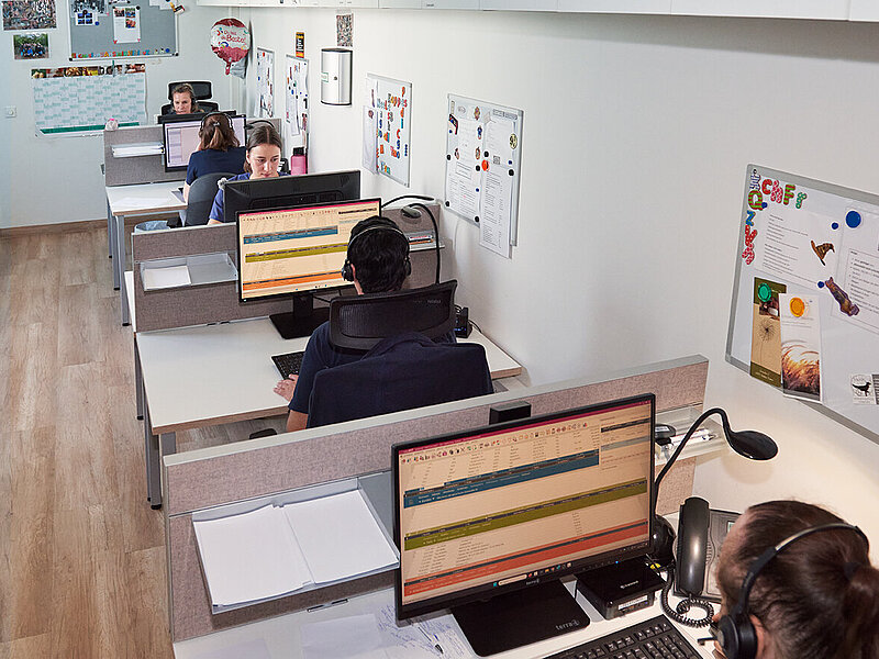 Das Bild zeigt unsere Telefonzentrale. Es sind fünf Mitarbeiterinnen zu sehen, die mit Headset telefonieren. Sie sitzen alle an kleinen Tischen vor Computerbildschirmen.