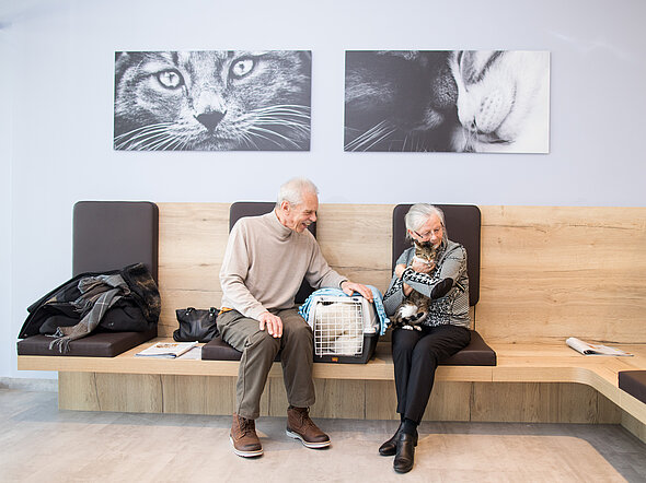 Hier sieht man zwei Katzenbesitzer:innen, die im Katzenwartezimmer entspannt mir ihrer Katze auf den Termin warten.