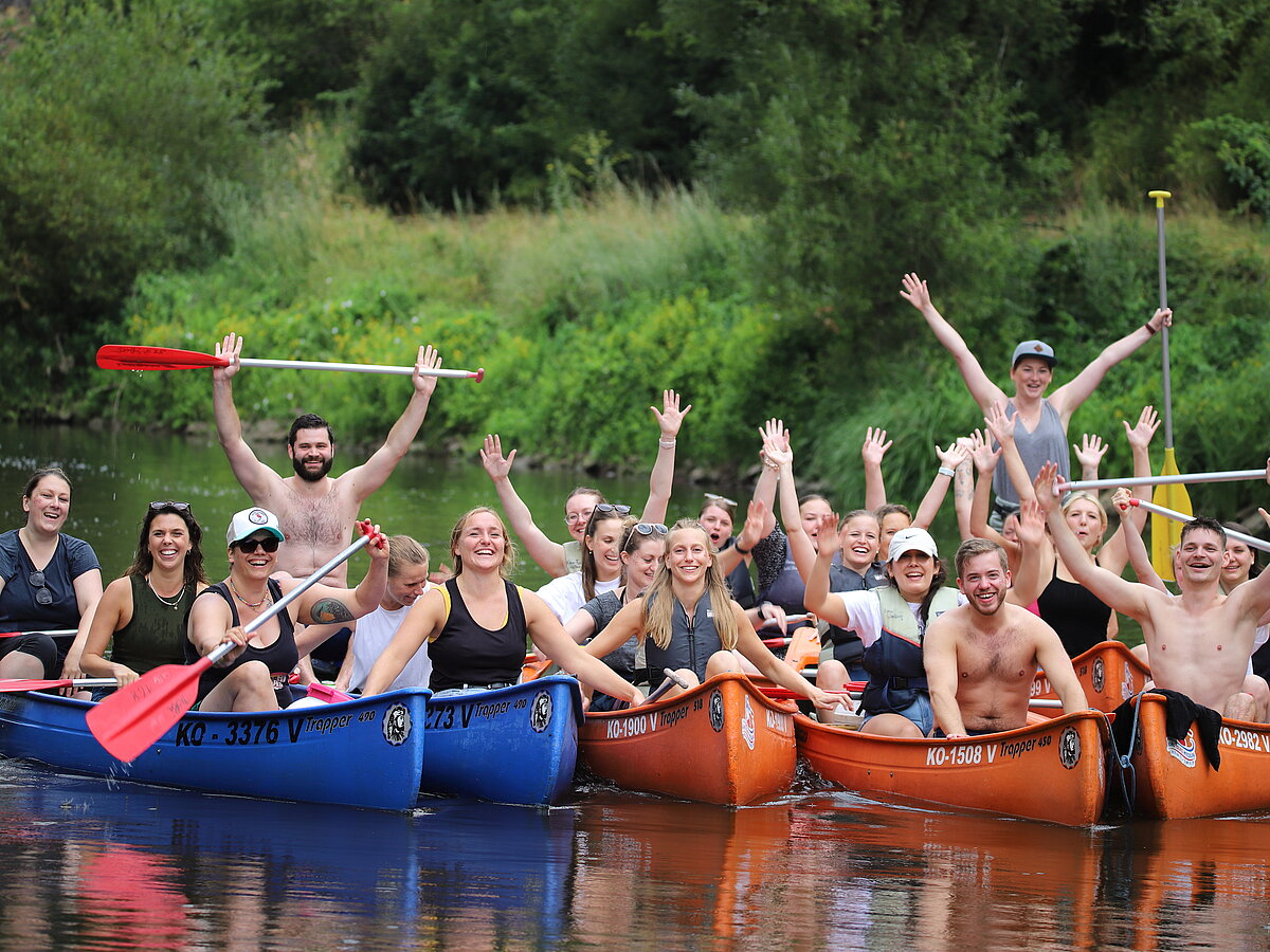 Hier sieht man mehrere Kanus auf einem Fluss, die voll mit Teammitgliedern beladen sind. Alle haben viel Spaß.